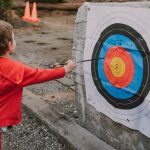 Young child in red hoodie practicing archery outdoors, successfully hitting the target.